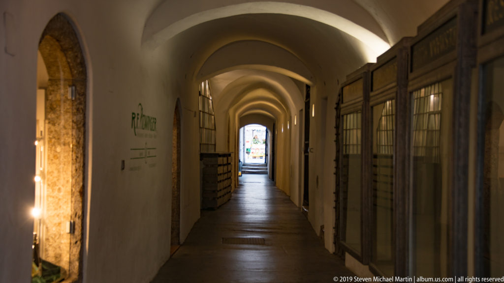 Long white vaulted passage lined with shopfronts, receding toward a bright doorway at the far end.