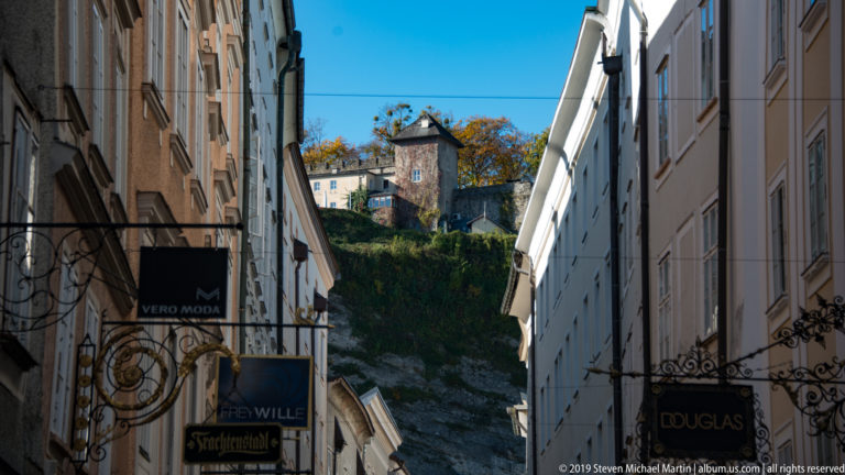 Narrow Getreidegasse lined with hanging shop signs and tall façades, ending at a rocky green hillside with a small tower.