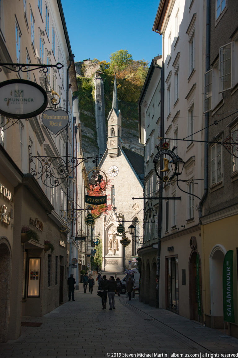 Narrow Getreidegasse with hanging shop signs leading toward St. Blasius Church and the steep green Mönchsberg slope.