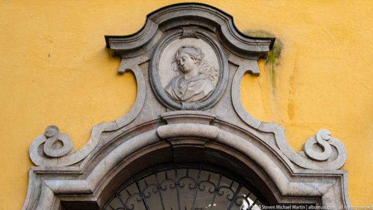 Oval stone portrait relief centered above a curved doorway surround with scroll-like volutes on a yellow wall.