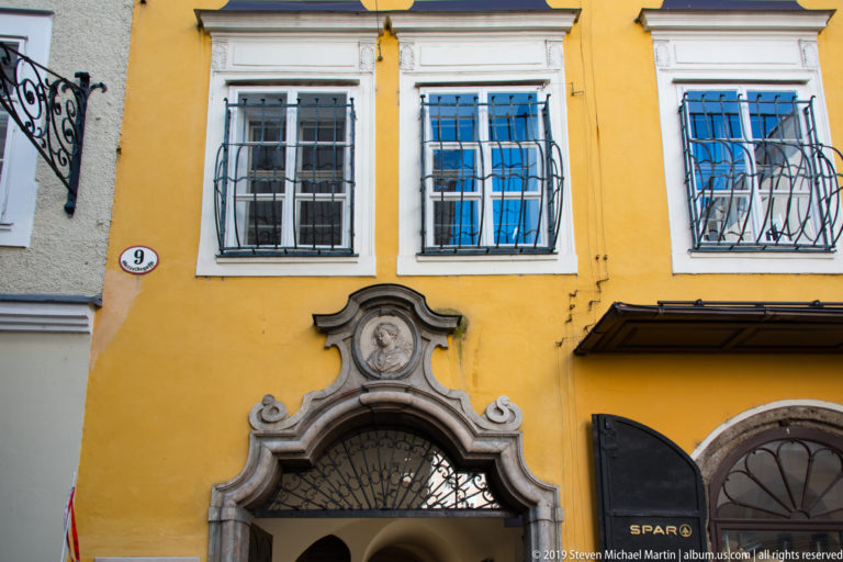 Ornate gray stone portal at Getreidegasse number 9 with portrait medallion, curved volutes and fan-shaped iron grille.