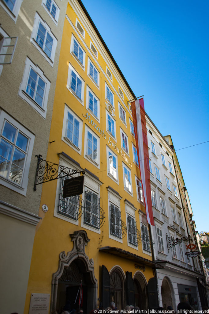 Tall yellow Getreidegasse façade at number 9 with white-trimmed windows, iron grilles, sculpted portal and Austrian flag.