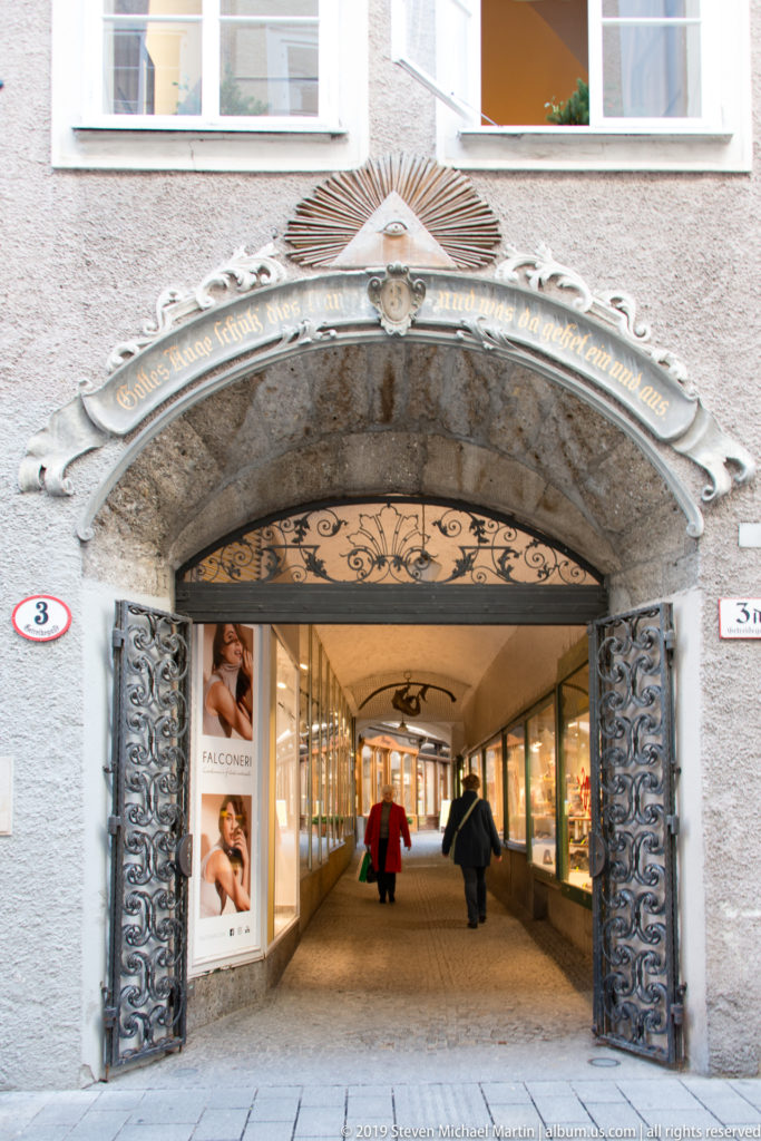 Stone-framed arched passage with decorative iron gates leading through a building to a vaulted shopping corridor.