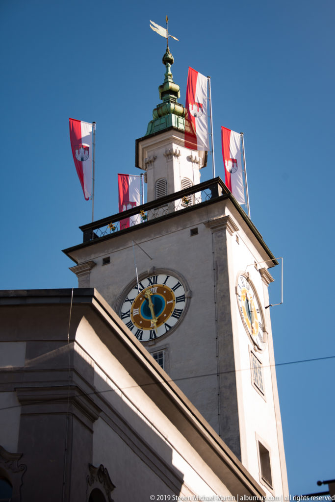 Close view of Salzburg Rathaus tower with two clock faces, a green roof crown and red-and-white flags against blue sky.