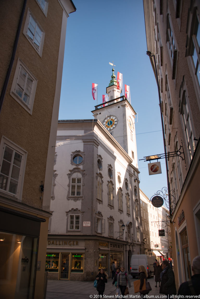 Salzburg Rathaus tower with clock faces and flags rising above a narrow street lined with historic buildings and shop signs.