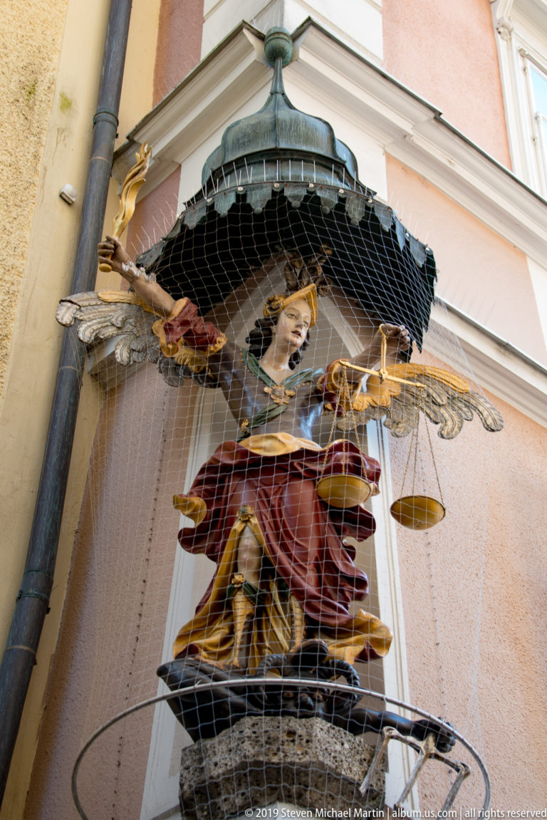 Polychrome winged figure holding balance scales beneath a small canopy, partly covered by protective netting.