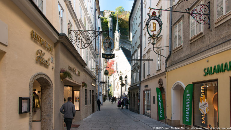Pedestrians walk along narrow Getreidegasse beneath projecting wrought-iron shop signs and closely spaced historic façades.