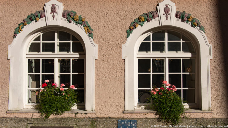 Two white-trimmed arched windows with flower boxes on a pink stucco façade decorated with small floral ornaments.