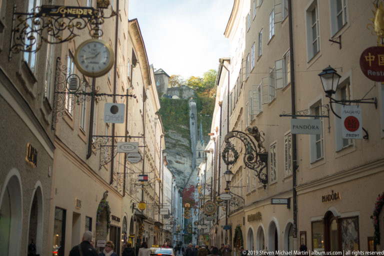 Narrow Getreidegasse lined with pale façades, hanging wrought-iron signs, pedestrians and a rocky slope in the distance.