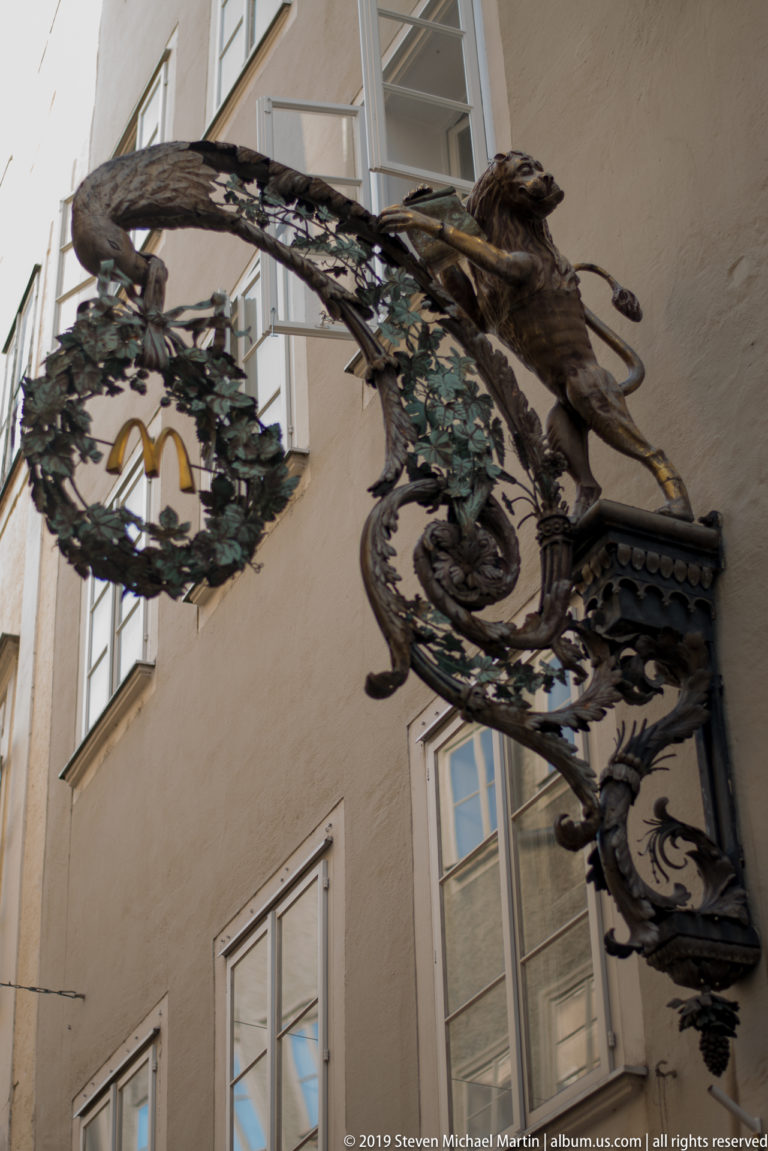 Ornate wrought-iron shop sign with leafy scrollwork and a gilded central emblem projecting from a pale façade.