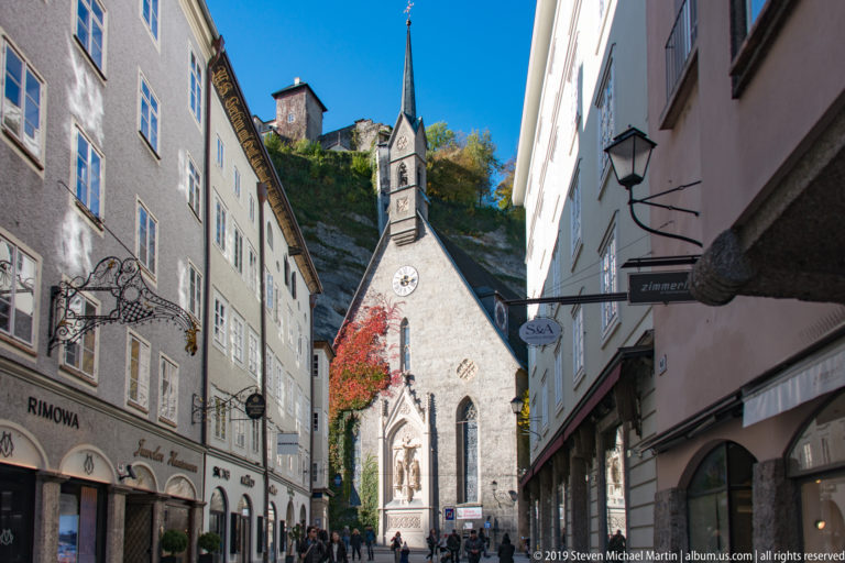 Getreidegasse leading toward the Gothic St. Blasius Church, with its slender turret and the Mönchsberg hillside rising behind.