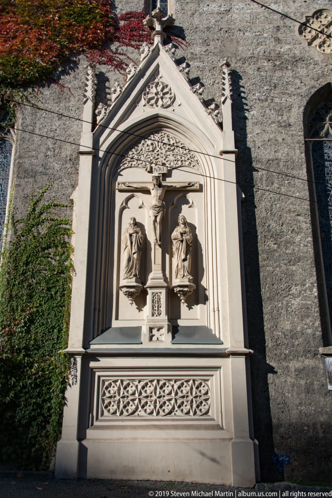 Pale stone Gothic-style wall monument with two robed figures beneath a pointed canopy and quatrefoil tracery.