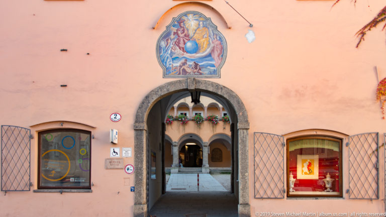 Stone-arched passage through a salmon façade beneath a colorful religious mural, opening into an arcaded courtyard.