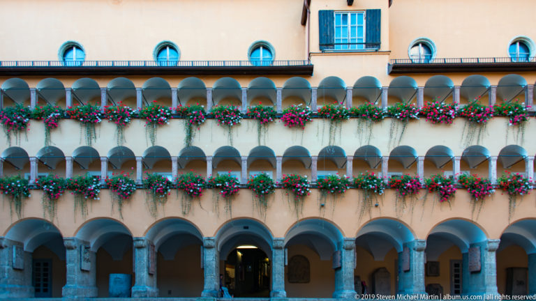 Pale courtyard façade with three levels of arcades and continuous flower boxes above a stone ground-floor colonnade.
