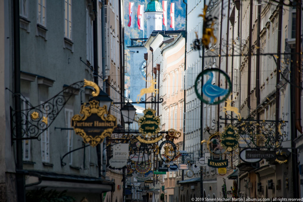 Layered wrought-iron business signs fill Getreidegasse, with a blue-green bird emblem prominent near the center.
