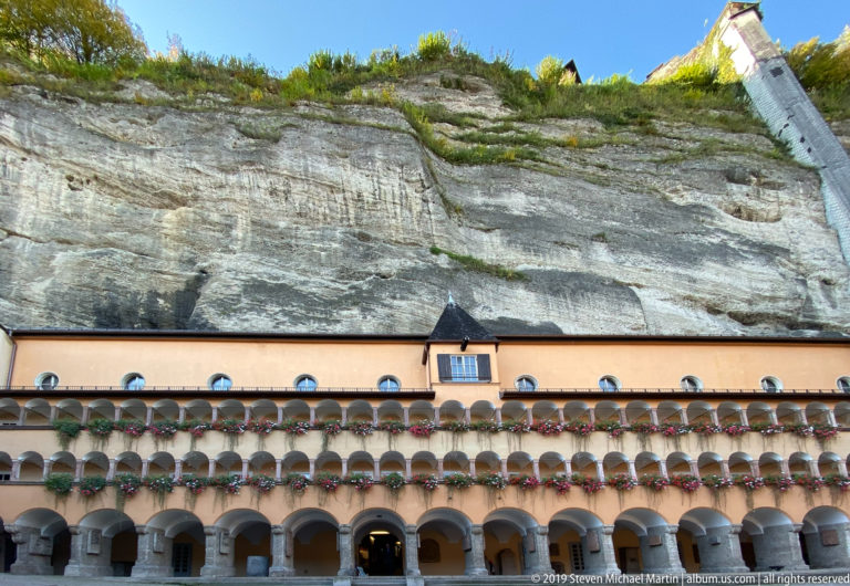 Long historic arcaded building with flower boxes directly beneath the sheer exposed rock face of the Mönchsberg.