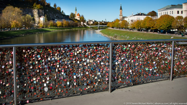 Colorful love locks covering a bridge railing, with the Salzach, church towers, and autumn-lined riverbanks beyond.