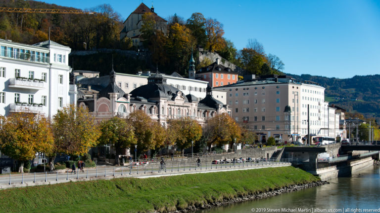 Historic pale buildings and autumn trees lining the Salzach, with wooded hillside and a hilltop structure above.