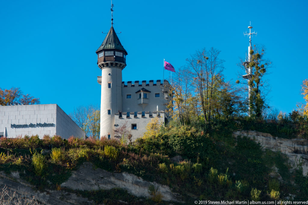 White cylindrical tower with crenellated wall, dark conical roof, red flag, and antennas on a wooded Salzburg slope.