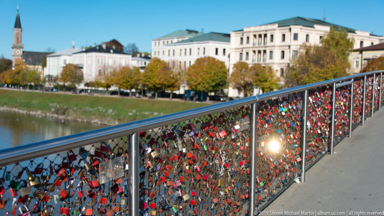 Bridge railing densely covered with colorful love locks, with Salzburg riverside buildings and autumn trees beyond.