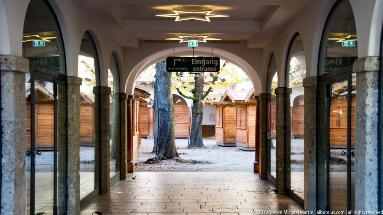 Arcaded passage with stone columns and an Eingang entrance sign opening toward wooden stalls beneath trees.