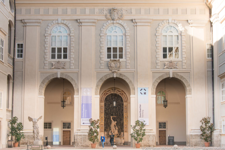 Pale Baroque courtyard façade with large arches, tall windows, sculptural masks, central decorated doorway, and potted trees.