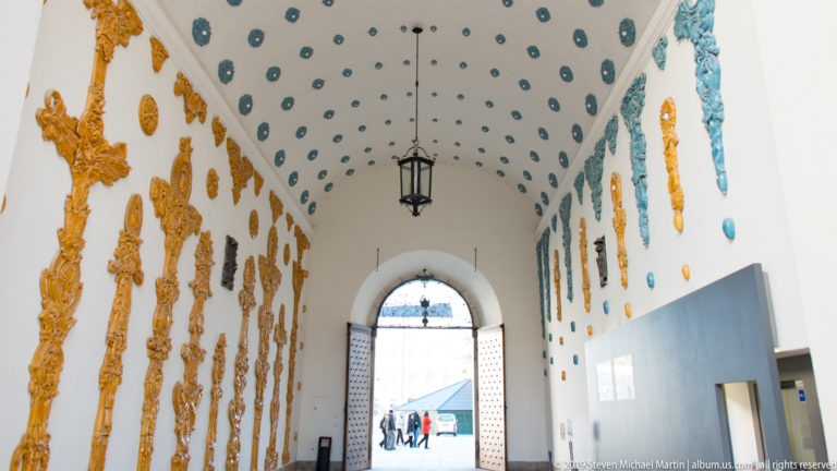 White vaulted passage decorated with blue-green rosettes, gilded reliefs, a hanging lantern, and an arched exit.