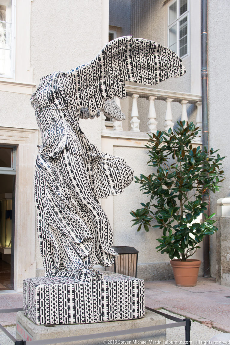 Large black-and-white patterned feline sculpture with long tail and crouching stance in a courtyard.