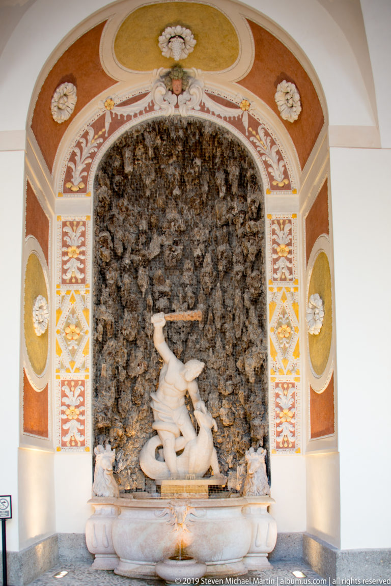 Classical reclining figure in a shallow fountain basin before a dark grotto niche framed by painted ornament.