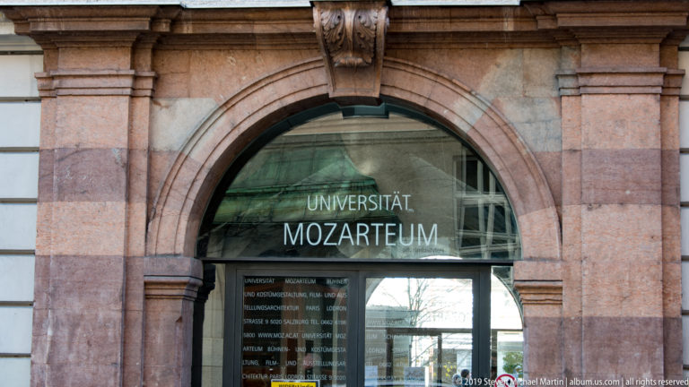 Arched stone entrance with glass doors labeled UNIVERSITÄT MOZARTEUM and reflections of the street beyond.