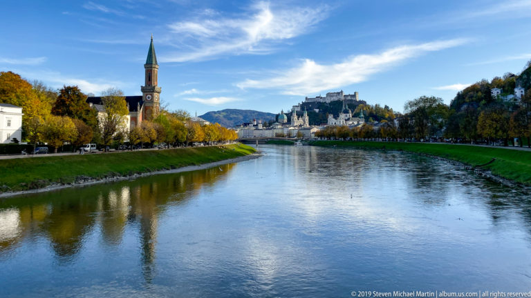 Wide Salzach River view with autumn trees, a church tower at left, and Hohensalzburg Fortress in the distance.