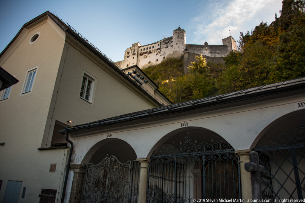 Hohensalzburg Fortress above autumn trees and low arcaded historic buildings with wrought-iron gates.
