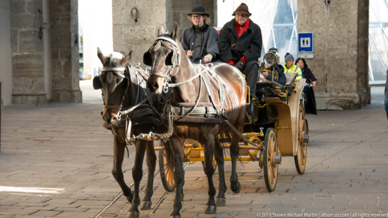Two dark horses pulling a traditional carriage with drivers and passengers through a stone-arched Salzburg passage.