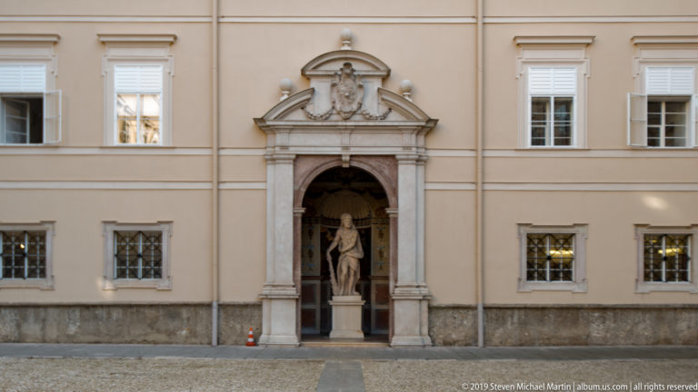 Ornate arched stone portal with sculptural cresting and a standing statue set into a pale symmetrical façade.