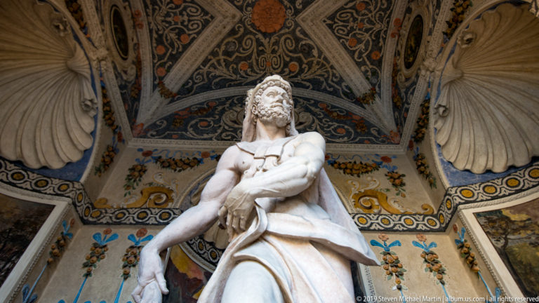 White marble male statue beneath an ornate painted vault with shells, arabesques, and colorful decorative panels.