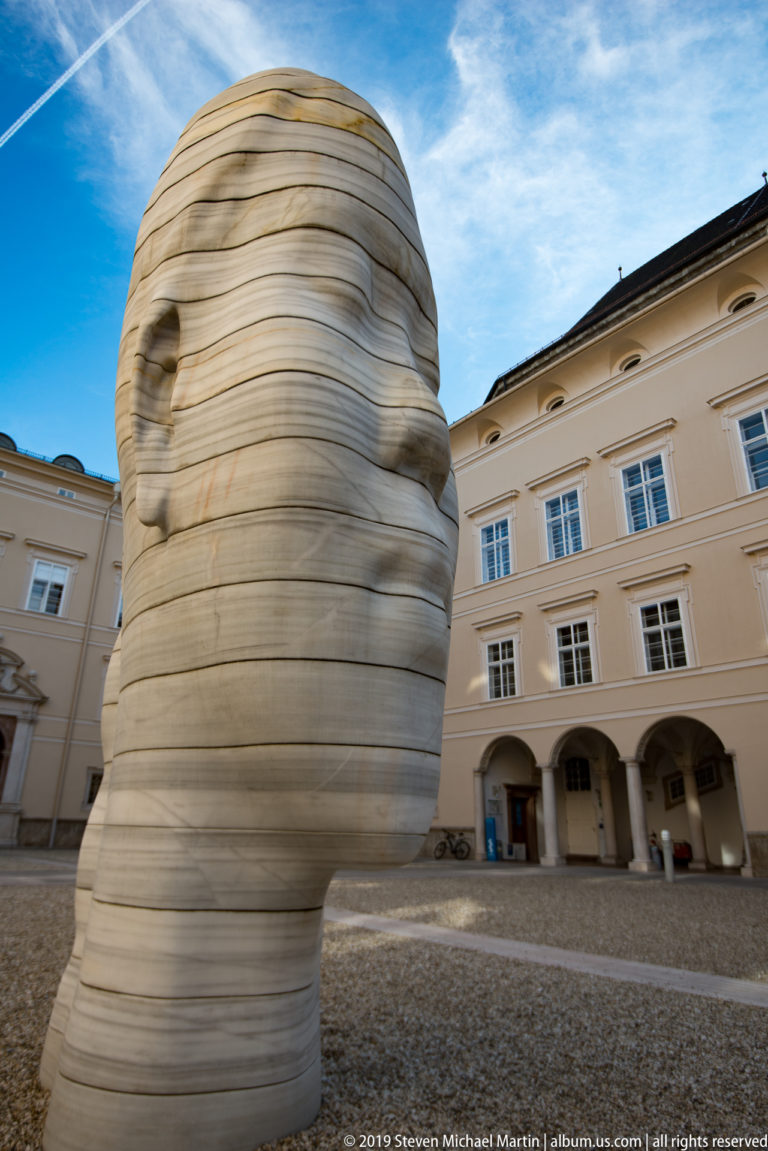 Large contemporary head sculpture made of horizontal layered bands, standing in a pale courtyard.