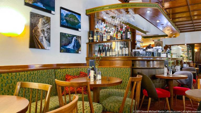 Colorful Salzburg café interior with patterned banquettes, wooden chairs, small tables, bar shelves, bottles, and hanging glasses.