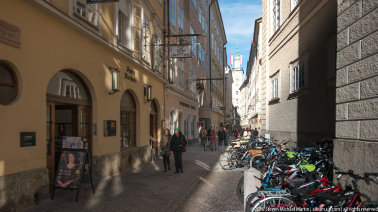 Narrow Salzburg pedestrian lane with historic façades, projecting shop signs, pedestrians, and bicycles parked along the right side.