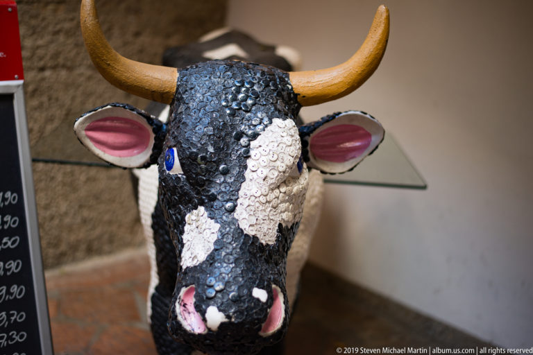 Sequined black-and-white cow sculpture with ochre horns, pink ears, blue eyes, and a bright pink muzzle.