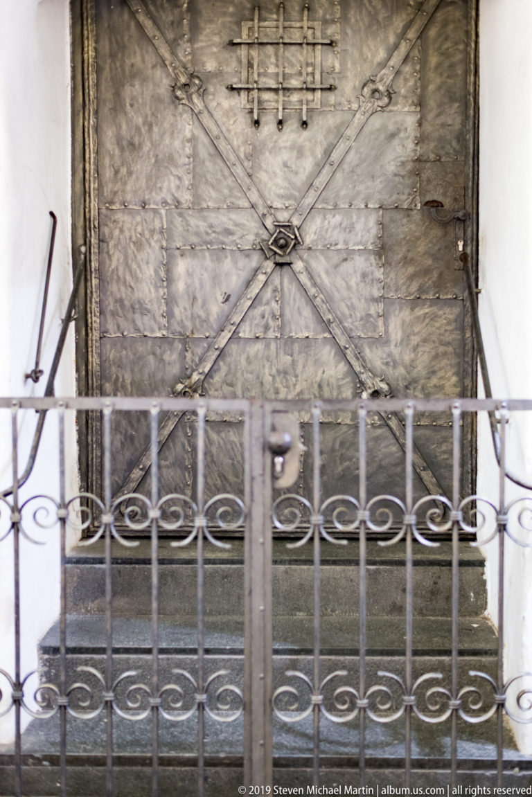 Weathered metal door with diagonal reinforcing bands and rivets, partly screened by a decorative iron railing.