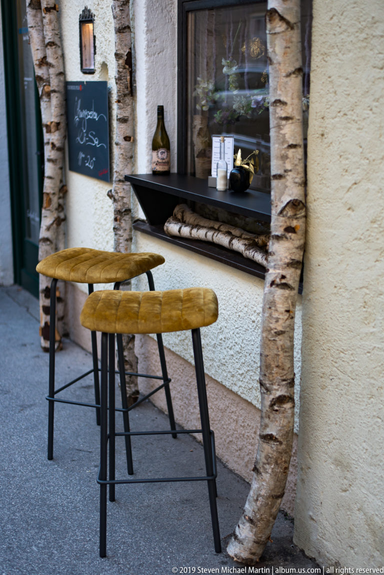 Two rustic wooden café tables beside a stone-framed service window with bottles and cups on the ledge.