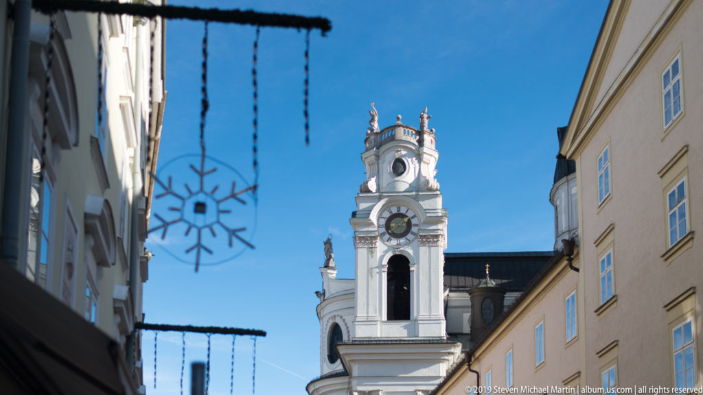 White Baroque church clock tower rising between narrow historic buildings, with hanging street decorations in the foreground.