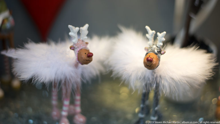 Two whimsical reindeer ornaments with antlers, striped legs, painted faces, and fluffy white feathered bodies.