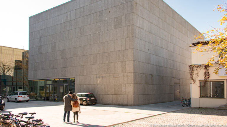 Modern pale-stone Universität Mozarteum building with low glass entrance, two people, bicycles, and autumn foliage.