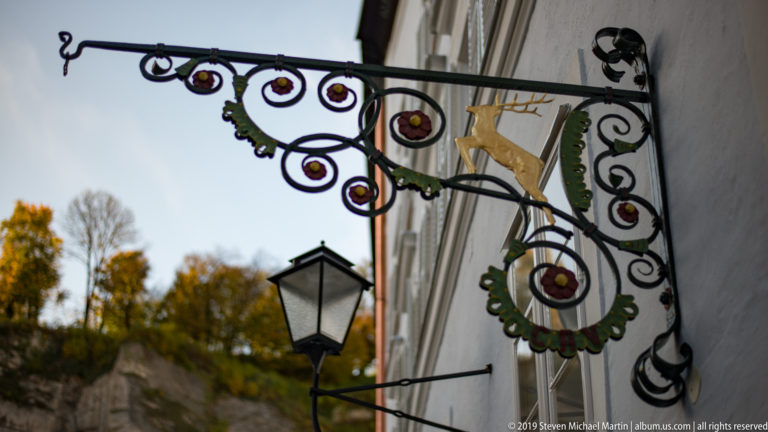 Decorative wrought-iron hanging sign with painted scrollwork and a small gilded figure beside a street lantern.