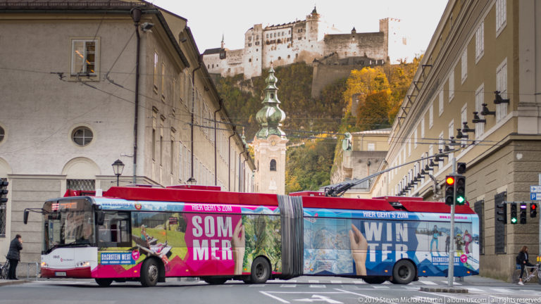 Colorful articulated city bus crossing a Salzburg street beneath Hohensalzburg Fortress and a green-domed church tower.