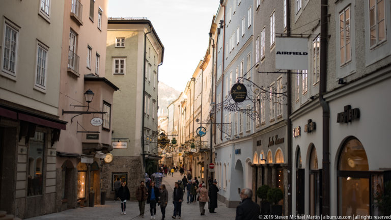 Pedestrians on narrow Getreidegasse beneath wrought-iron shop signs and historic façades, with mountains visible beyond.
