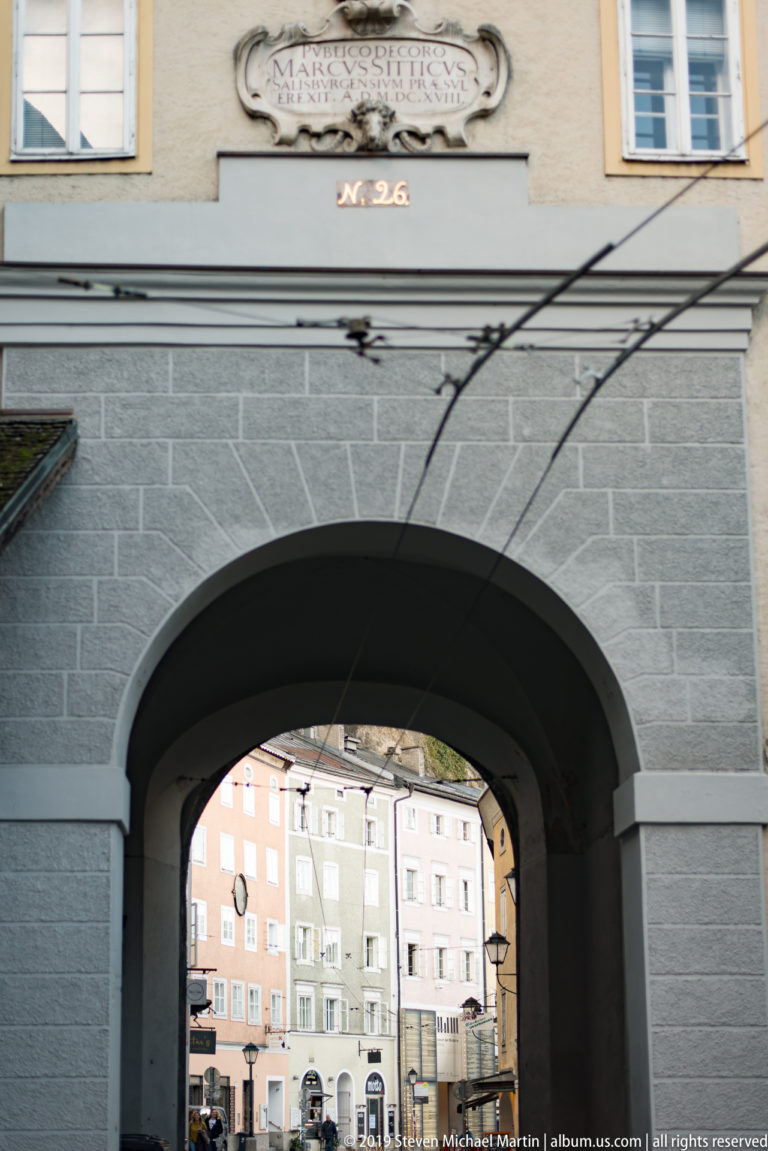 Large gray masonry archway framing pastel buildings and shops in a sunlit Salzburg square beyond.