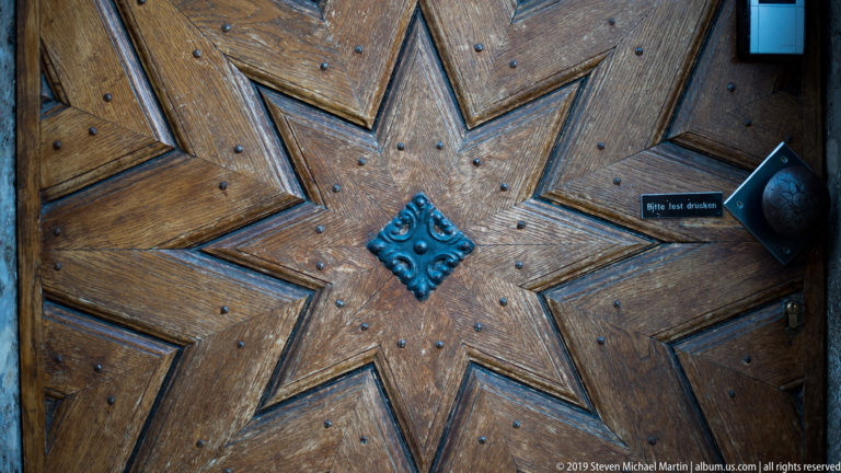 Close view of a heavy wooden door with geometric panels, iron studs, central metal boss, and a small German instruction sign.