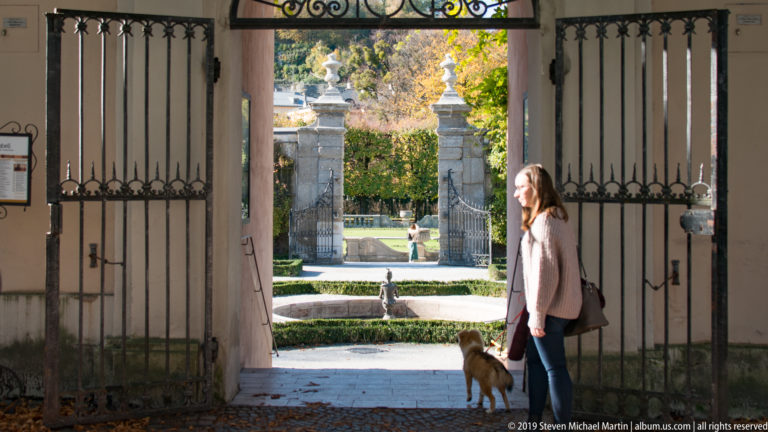 Woman standing with a small dog beneath open wrought-iron gates leading into a sunlit formal garden.